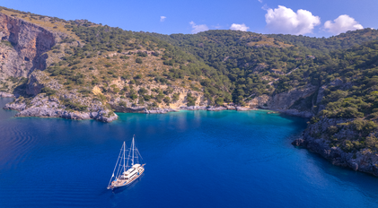 Aerial view of a forested Mediterranean peninsula opening into a secluded bay with a sailing yacht