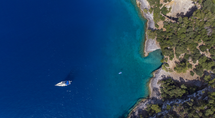 Aerial view of yacht drifting on deep blue waters beside forested rocky coastline