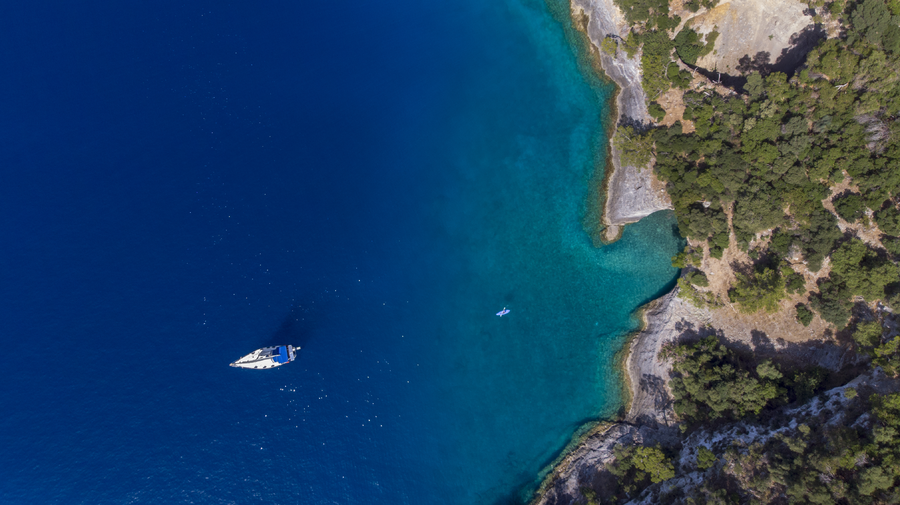 Aerial view of yacht drifting on deep blue waters beside forested rocky coastline