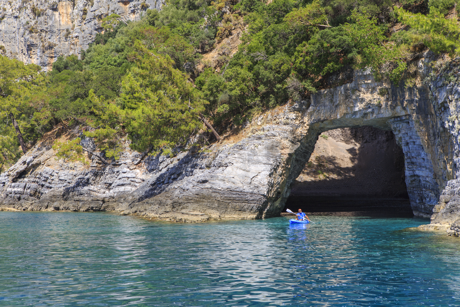 Kayaker entering a secluded forest-framed coastal arch on a Mediterranean peninsula