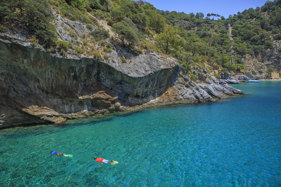 Snorkelers exploring a sheltered turquoise cove beneath rugged Mediterranean cliffs